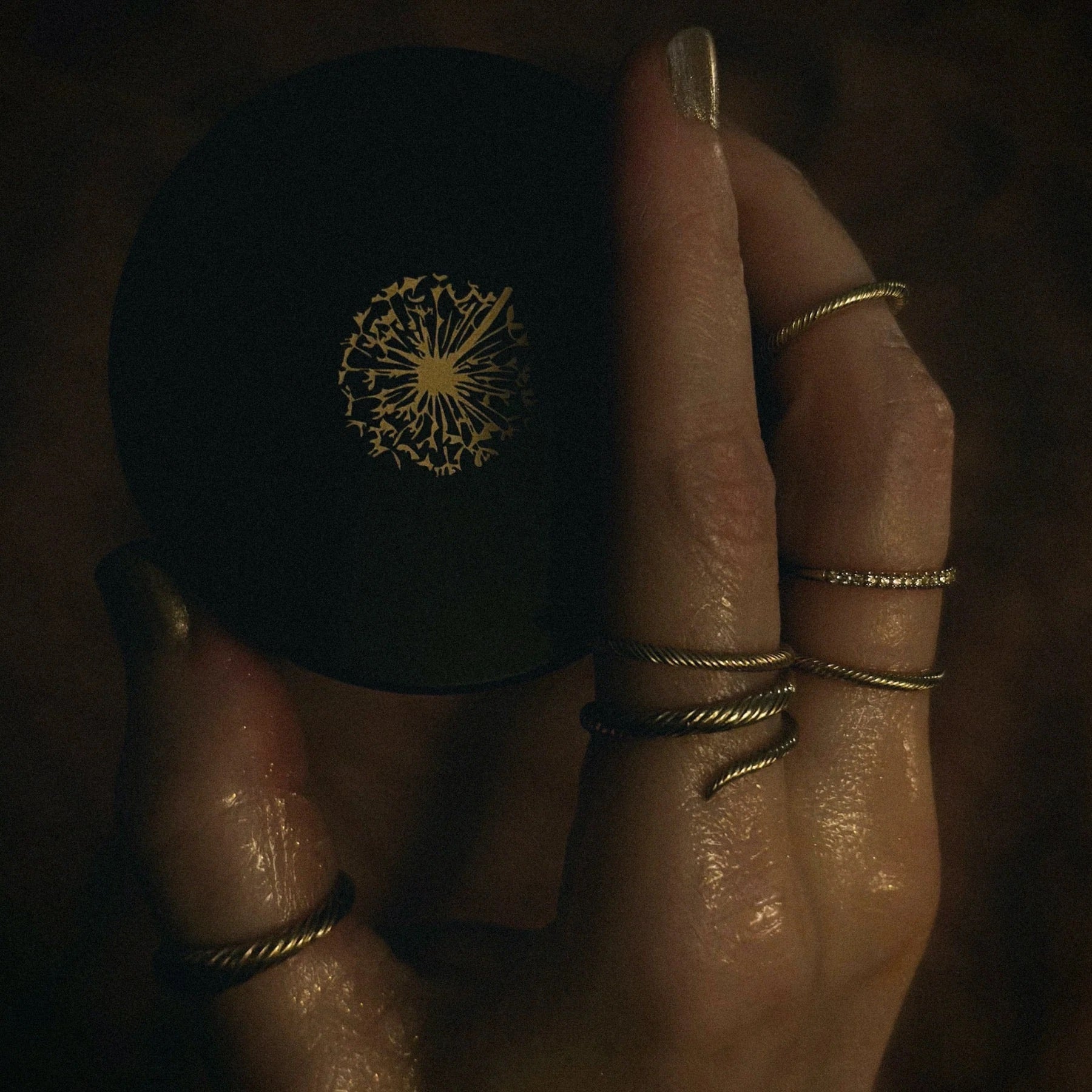 Hand covered in shimmering oil, wearing solid gold rings with twisted texture and diamonds, holding jar with dandelion logo.