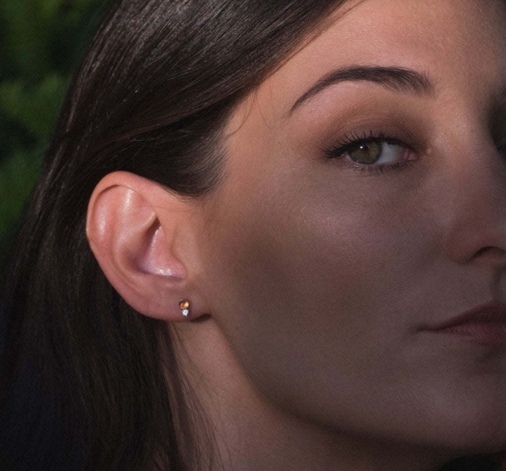 Solid gold gemstone earrings in glowing sunstone and white sapphire, worn by model with lush foliage in background.