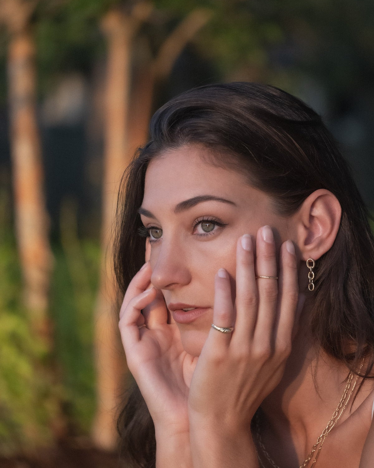 Woman wearing Aiden Jae textured yellow gold earrings, rings, and necklaces with trees in background.