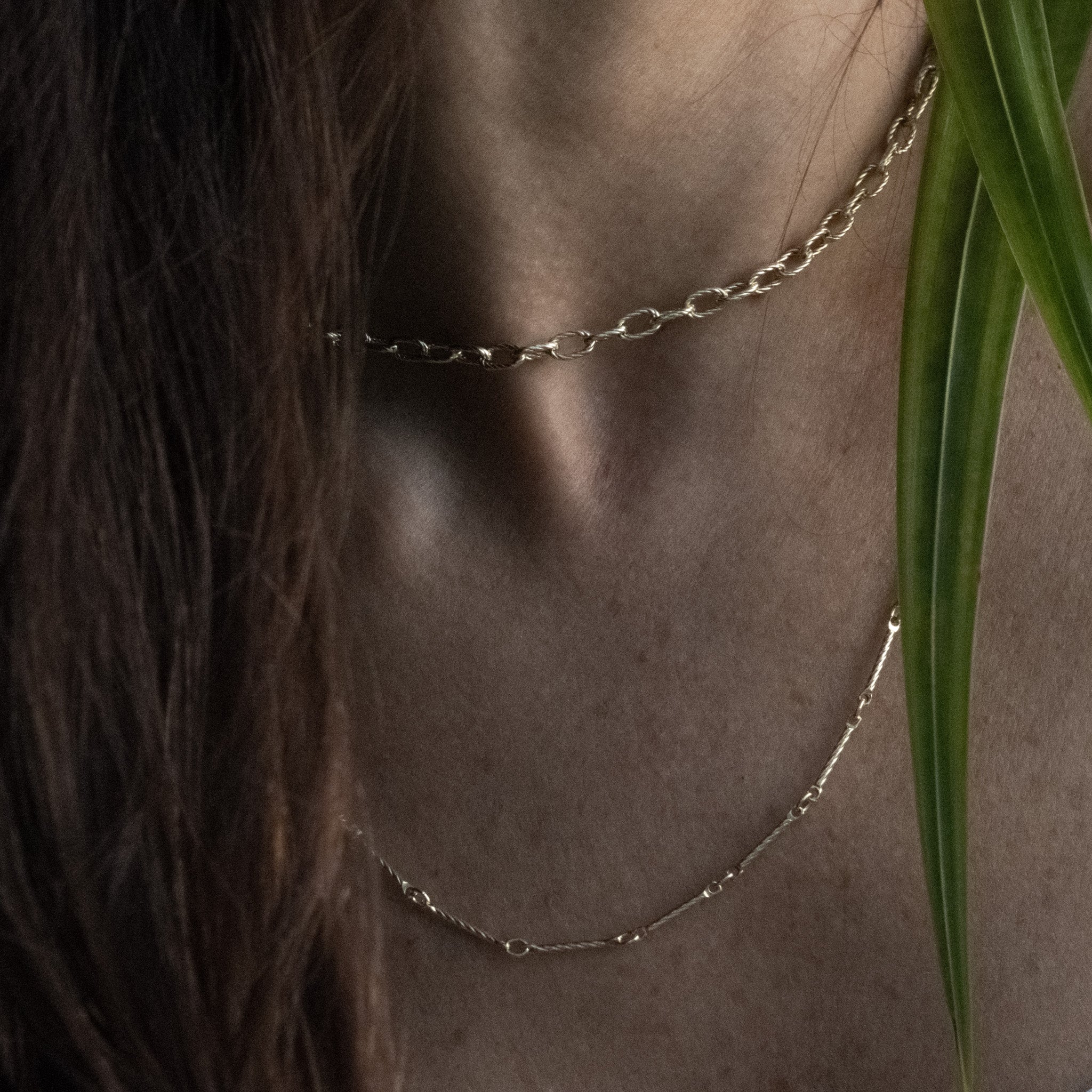 Solid gold cable and bar chain necklaces with twisted texture worn layered on neck with palm frond in foreground.