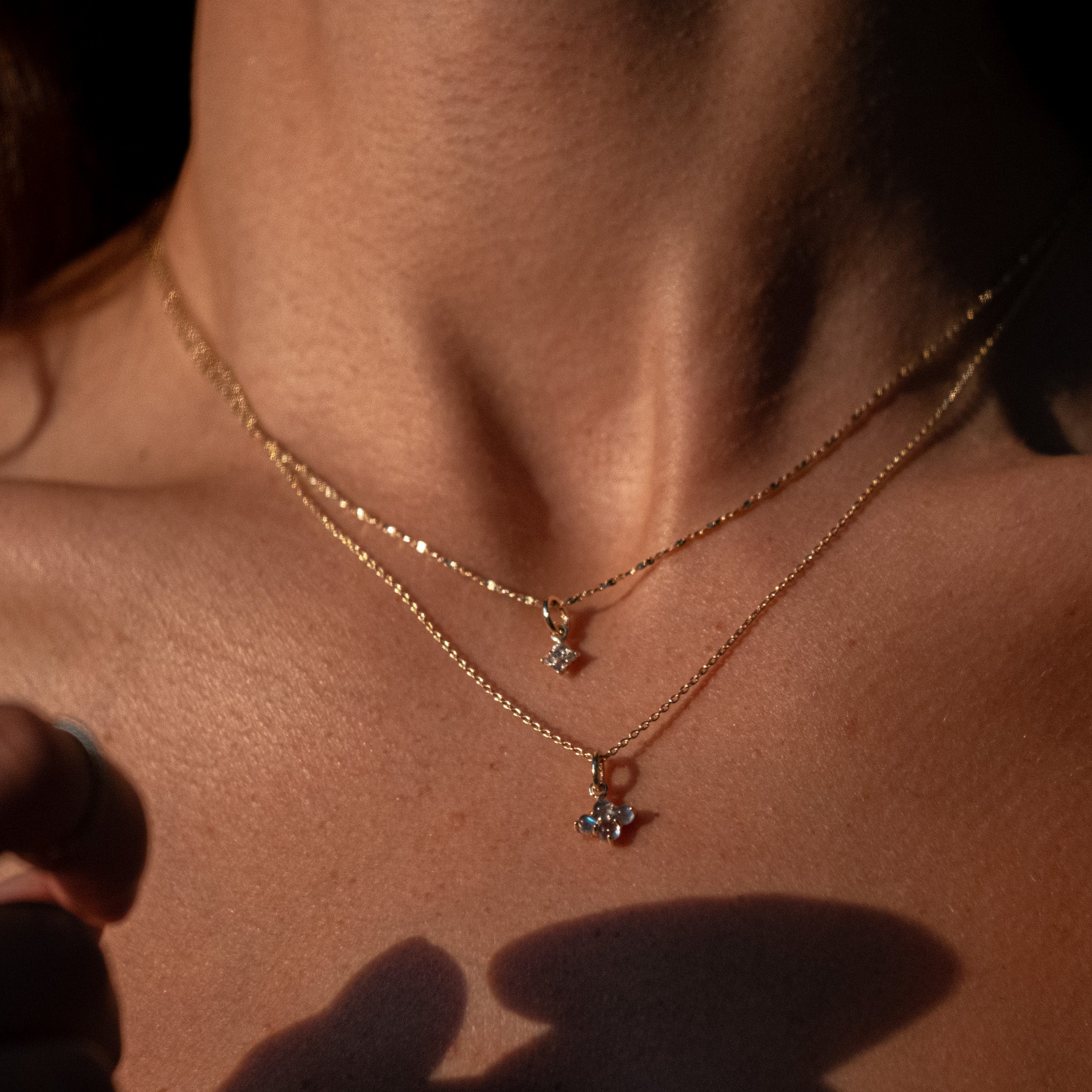 Solid gold white sapphire and glowing labradorite charm pendants on sparkling chain necklaces, layered on neck in sunlight.