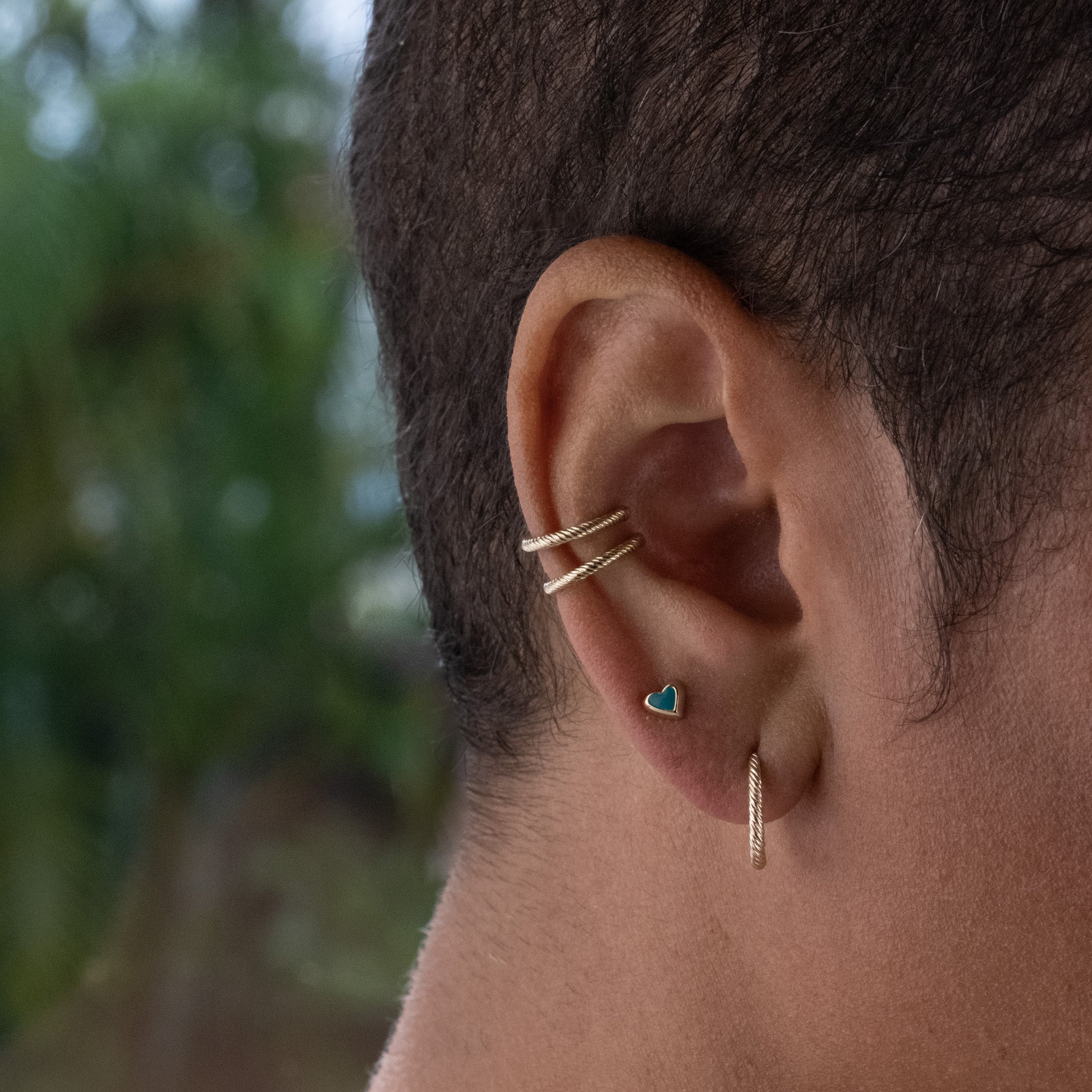 Solid gold tiny heart stud earring in teal enamel, with hoop and doubled ear cuffs in twisted texture, worn stacked on ear.