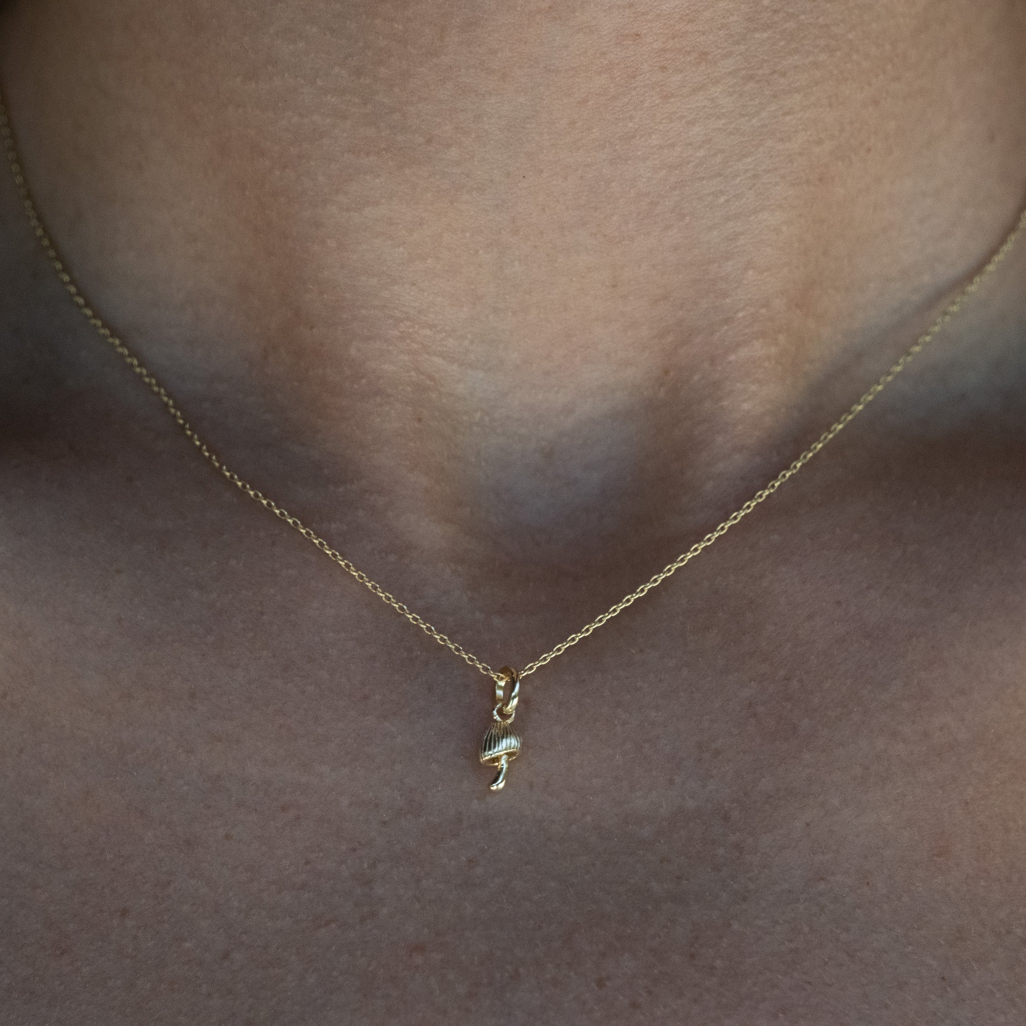 Solid gold tiny mushroom charm pendant, in a minimalist design, hanging on cable chain necklace, worn on neck.