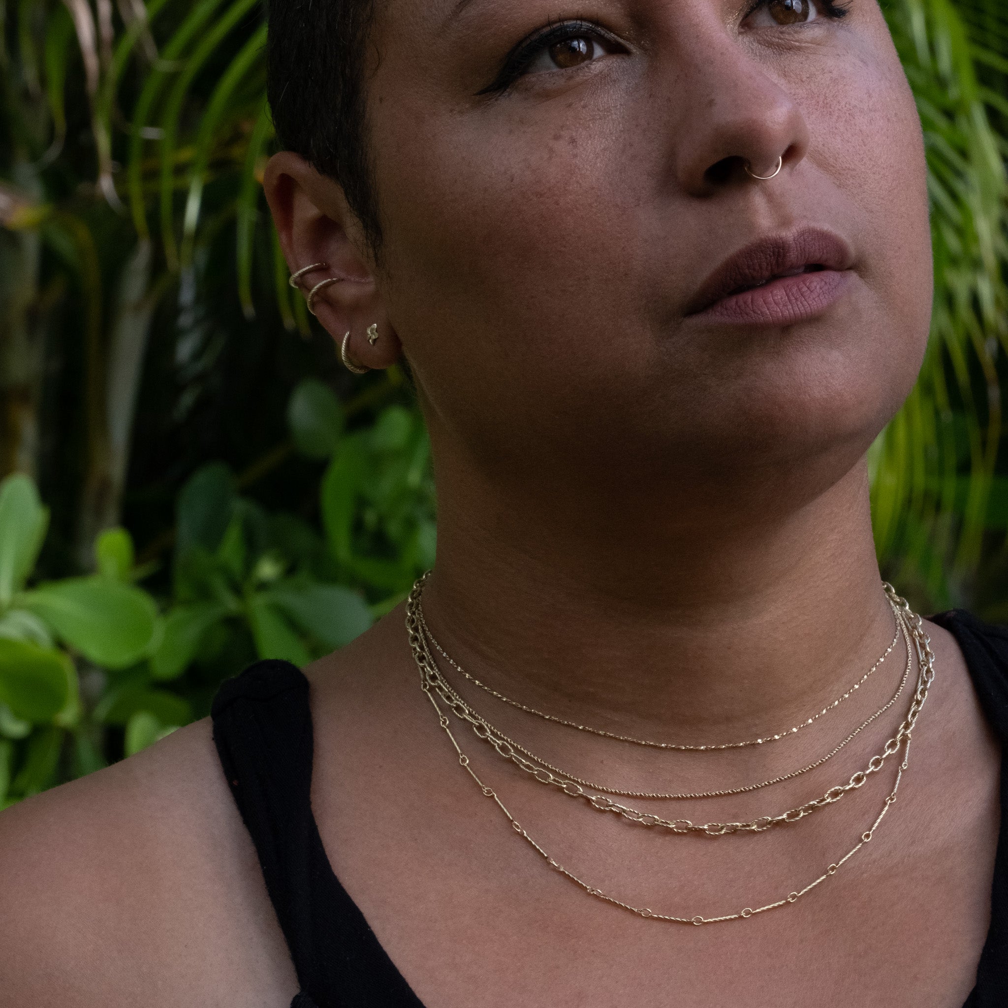 Solid gold twisted serpentine chain necklace styled with layered gold chains and earrings, tropical foliage in background.