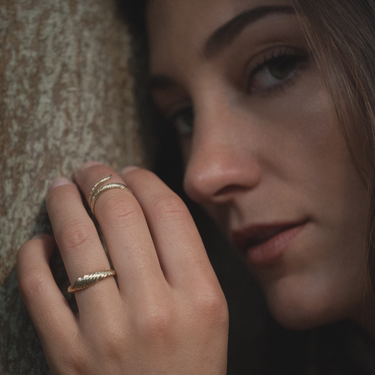 Model leaning on tree with hand near mouth wearing yellow gold rings with twisted texture.