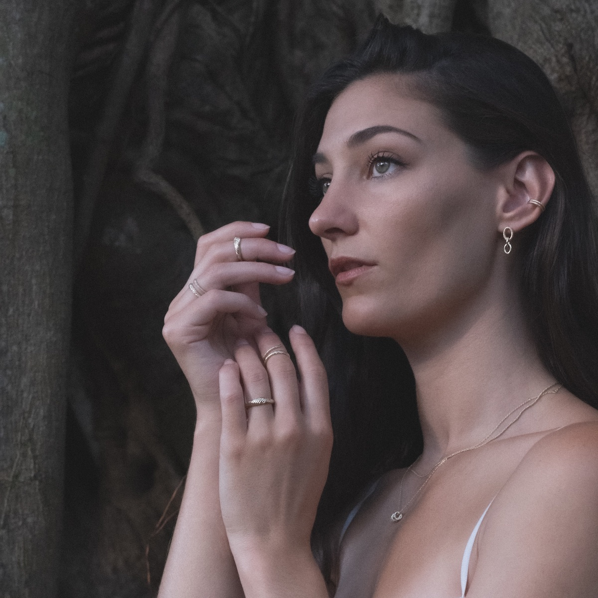 Model wearing yellow gold link earrings, ear cuffs, rings, and chains with twisted texture next to Banyan tree.