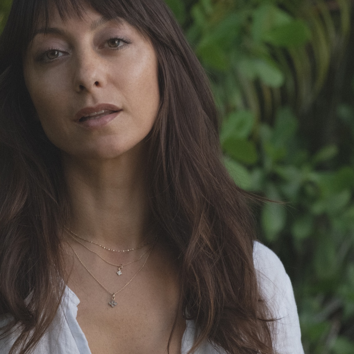 Model in white shirt in front of foliage wearing delicate yellow gold chains and small pendant charms in white sapphire and labradorite.