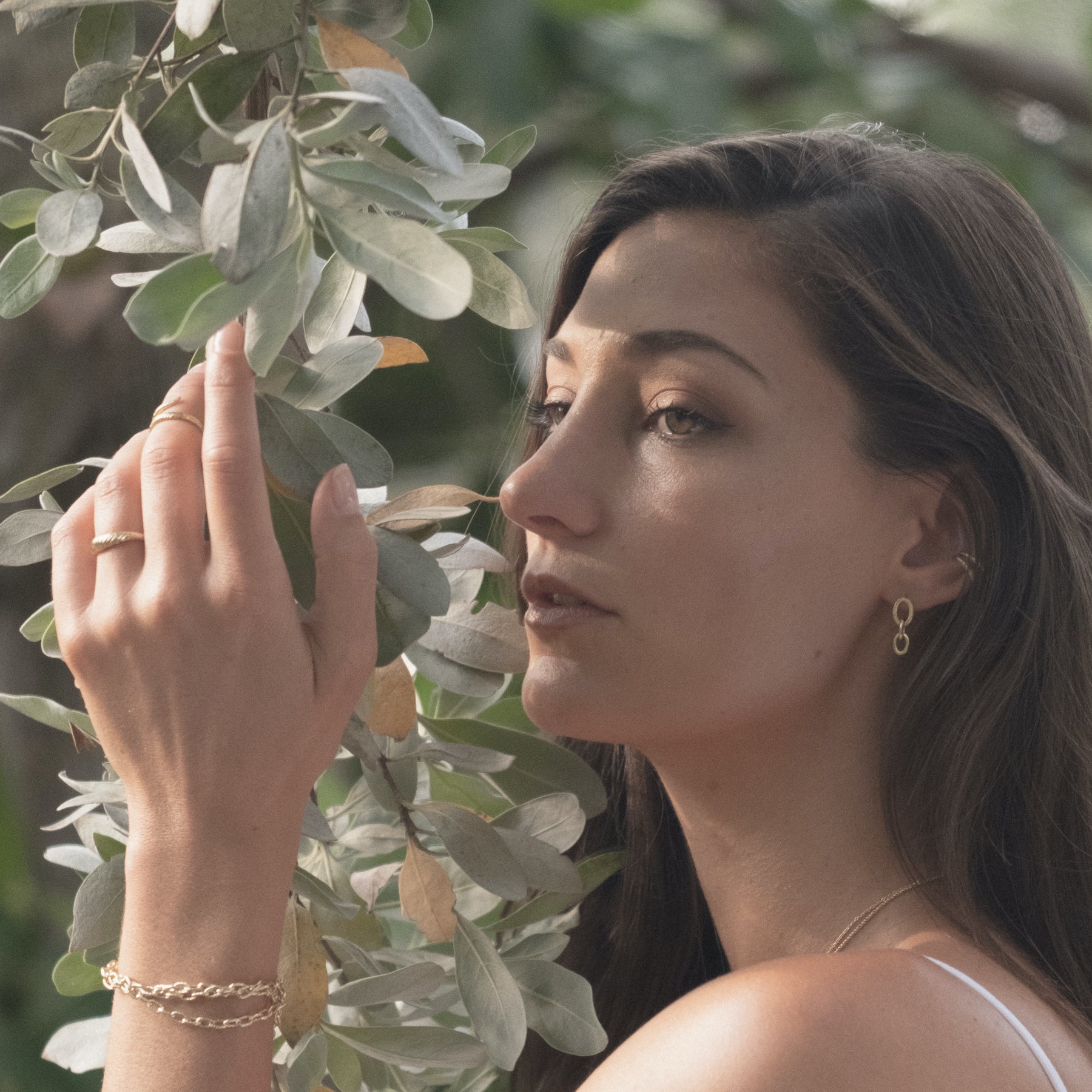 Model wearing textured, yellow gold rings, earrings, and bracelets from Aiden Jae. and holding a branch with green leaves. 