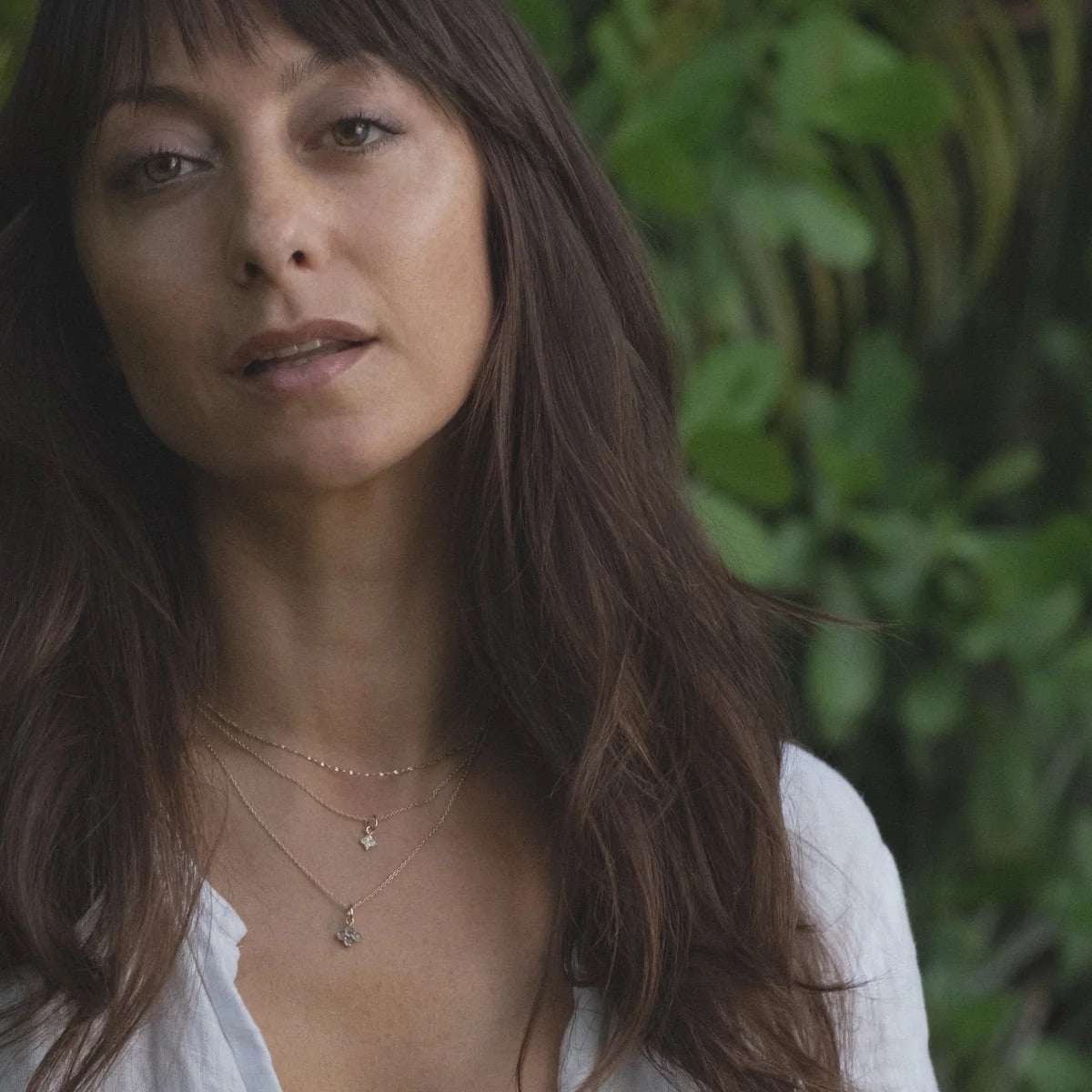 Solid gold and gemstone charm pendants on chains, layered on model with tropical foliage in background.