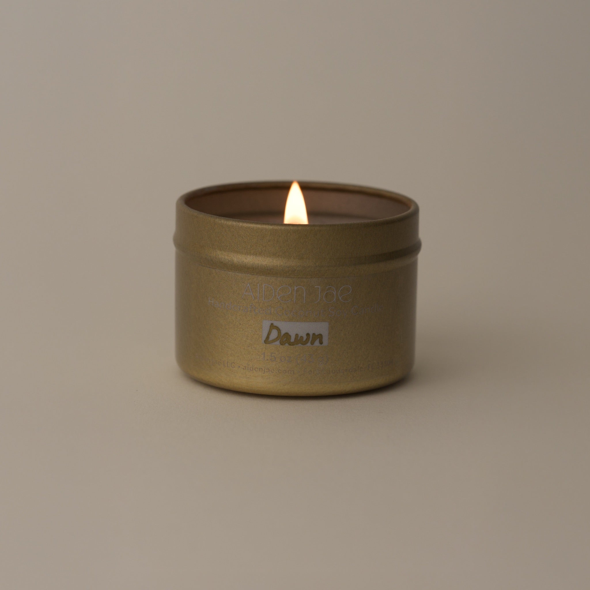 Mini candle in a gold tin, lit with visible flame, displayed against softly dimmed background.