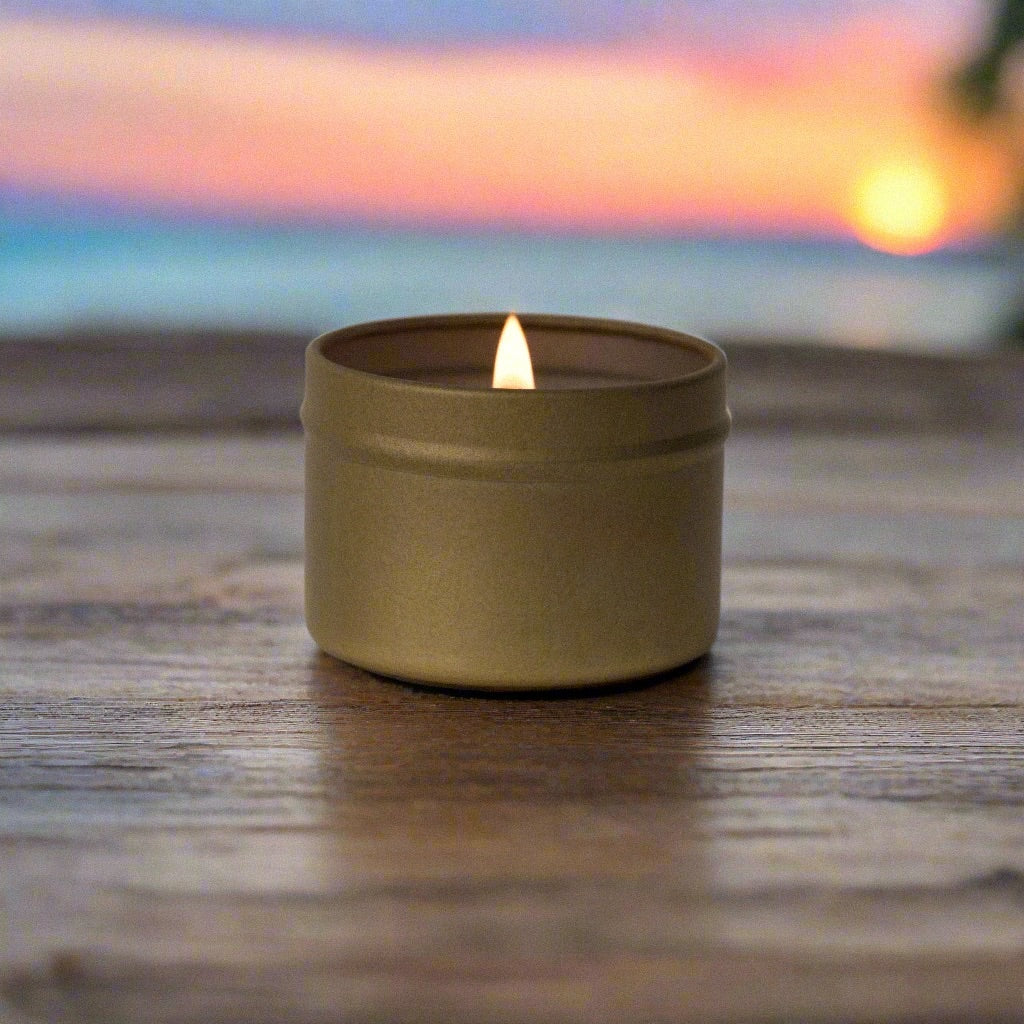Candle in a gold tin, lit with visible flame, placed on wood table with a tropical sky and beach shoreline in the background.
