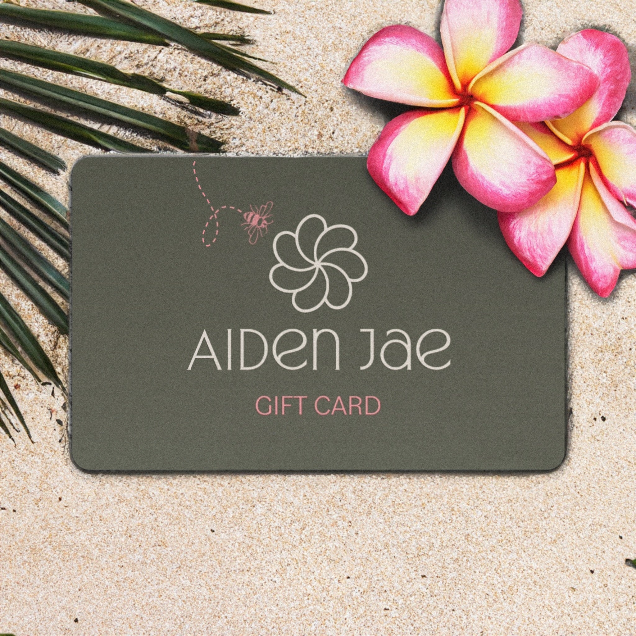 Aiden Jae digital gift card displayed in sand with plumeria flowers and a palm frond.