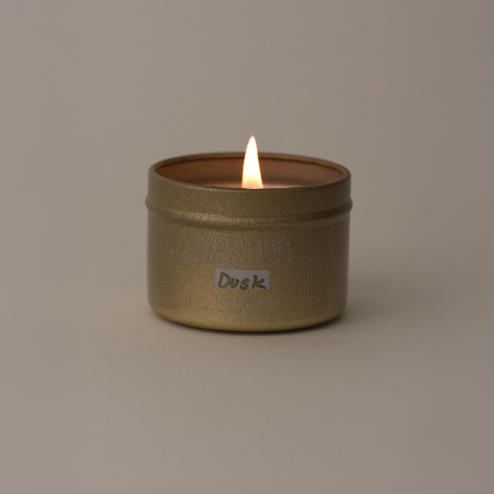 Mini candle in a gold tin, lit with visible flame, displayed against softly dimmed background.