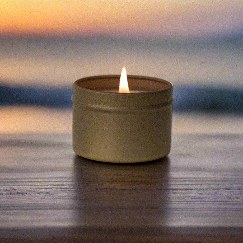 Candle in a gold tin, lit with visible flame, placed on wood table with a tropical sunset and beach in the background.