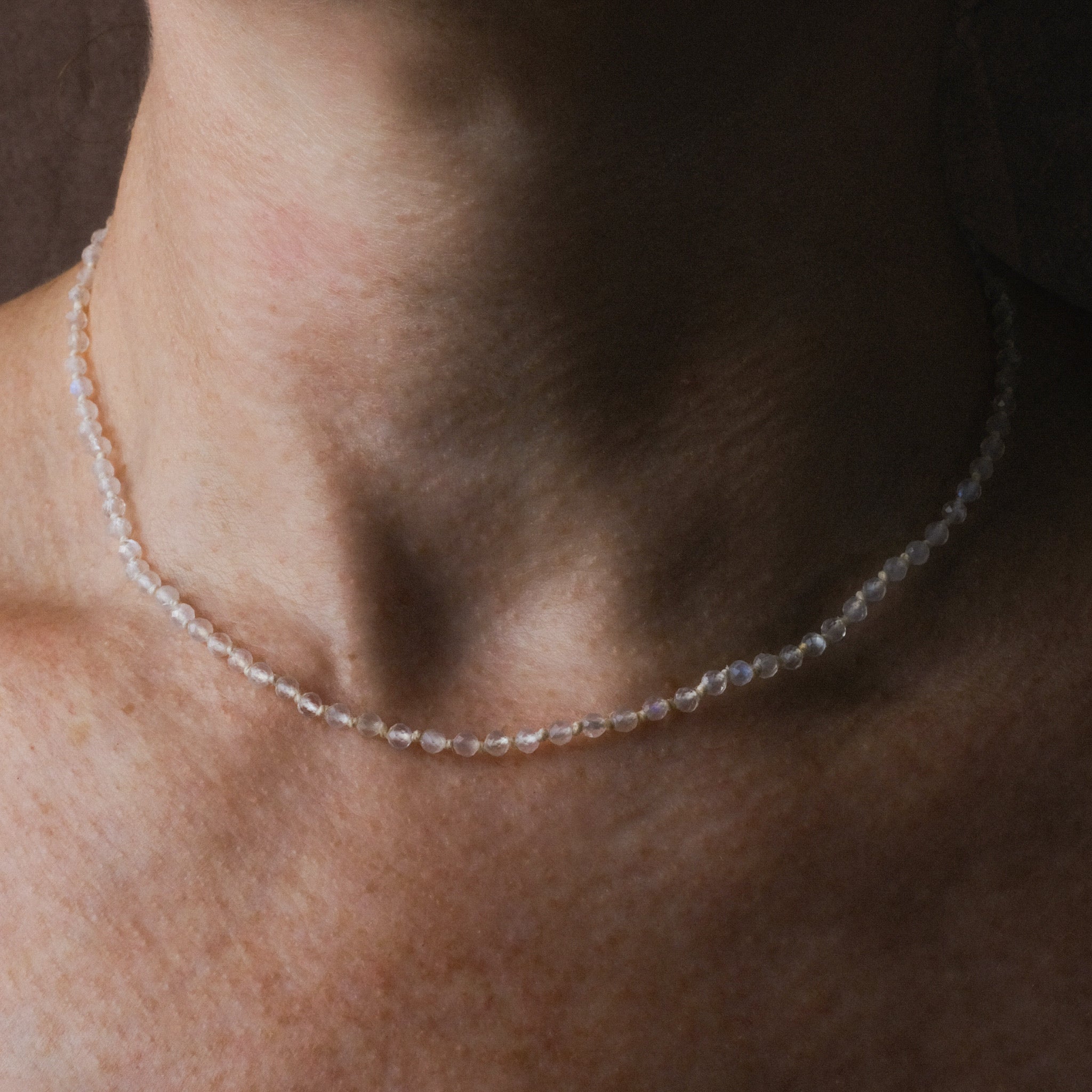 Knotted bead necklace with faceted rainbow moonstone and white hemp, worn on neck.