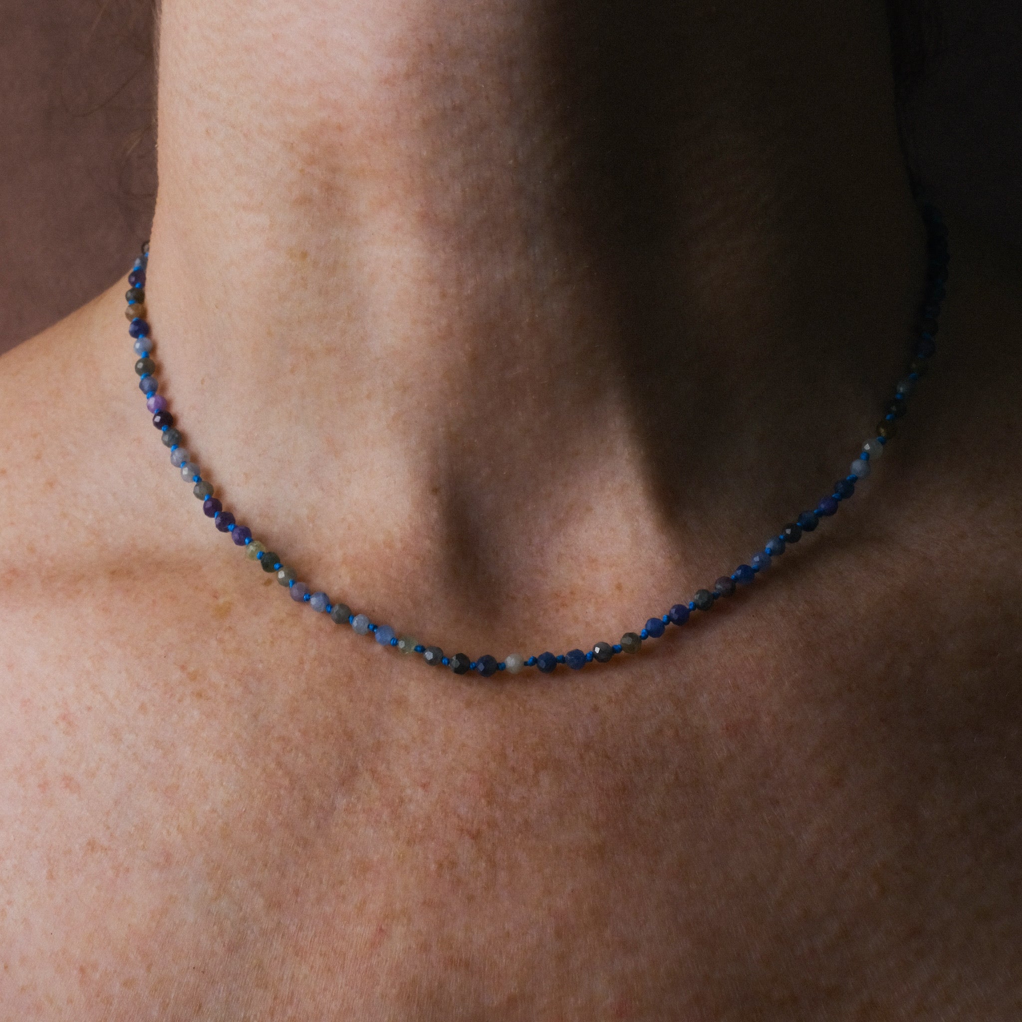 Knotted bead necklace with faceted multi-colored sapphire beads and bright blue hemp, worn on neck.