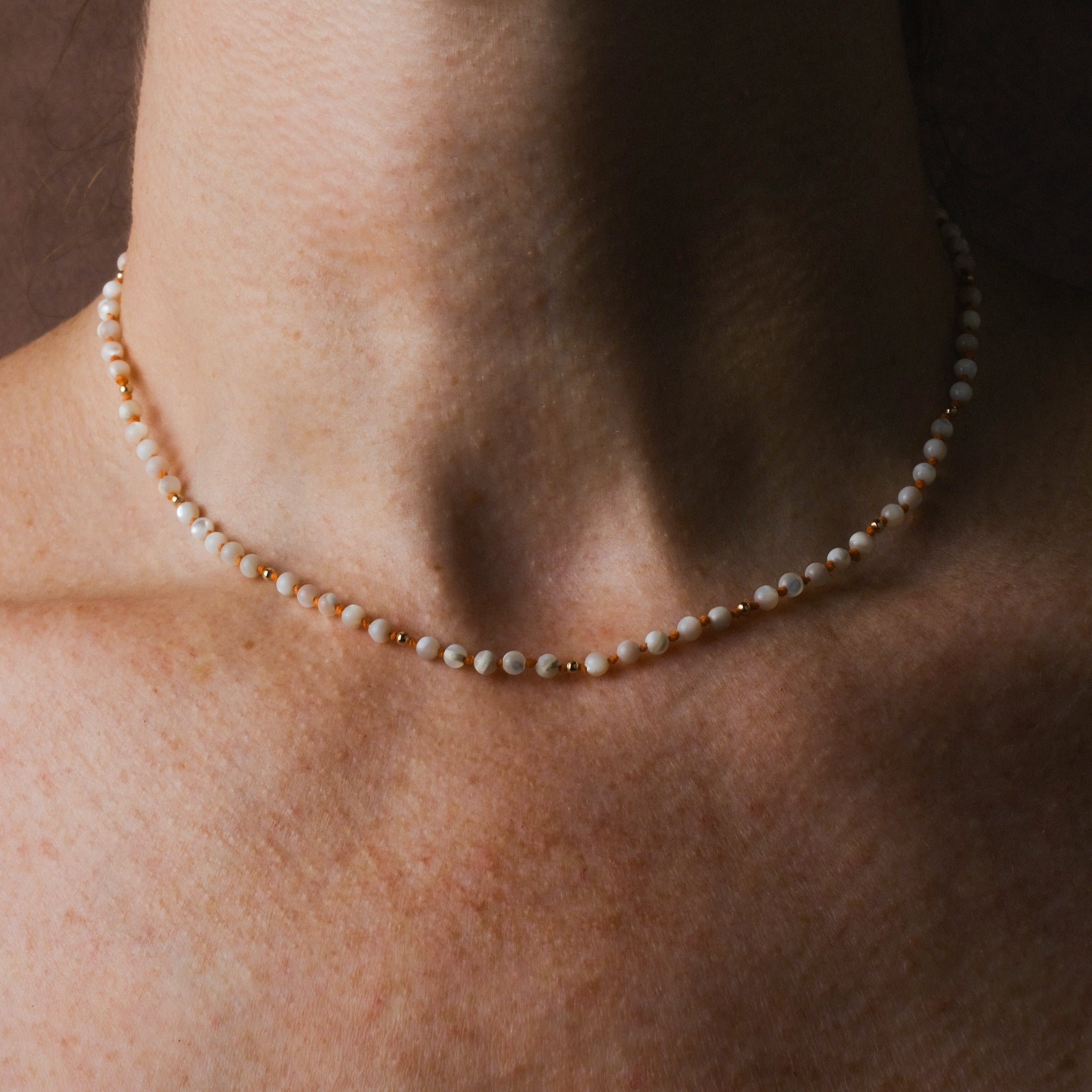 Knotted bead necklace with smooth mother-of-pearl, tiny solid gold beads, and bright orange hemp, worn on neck.