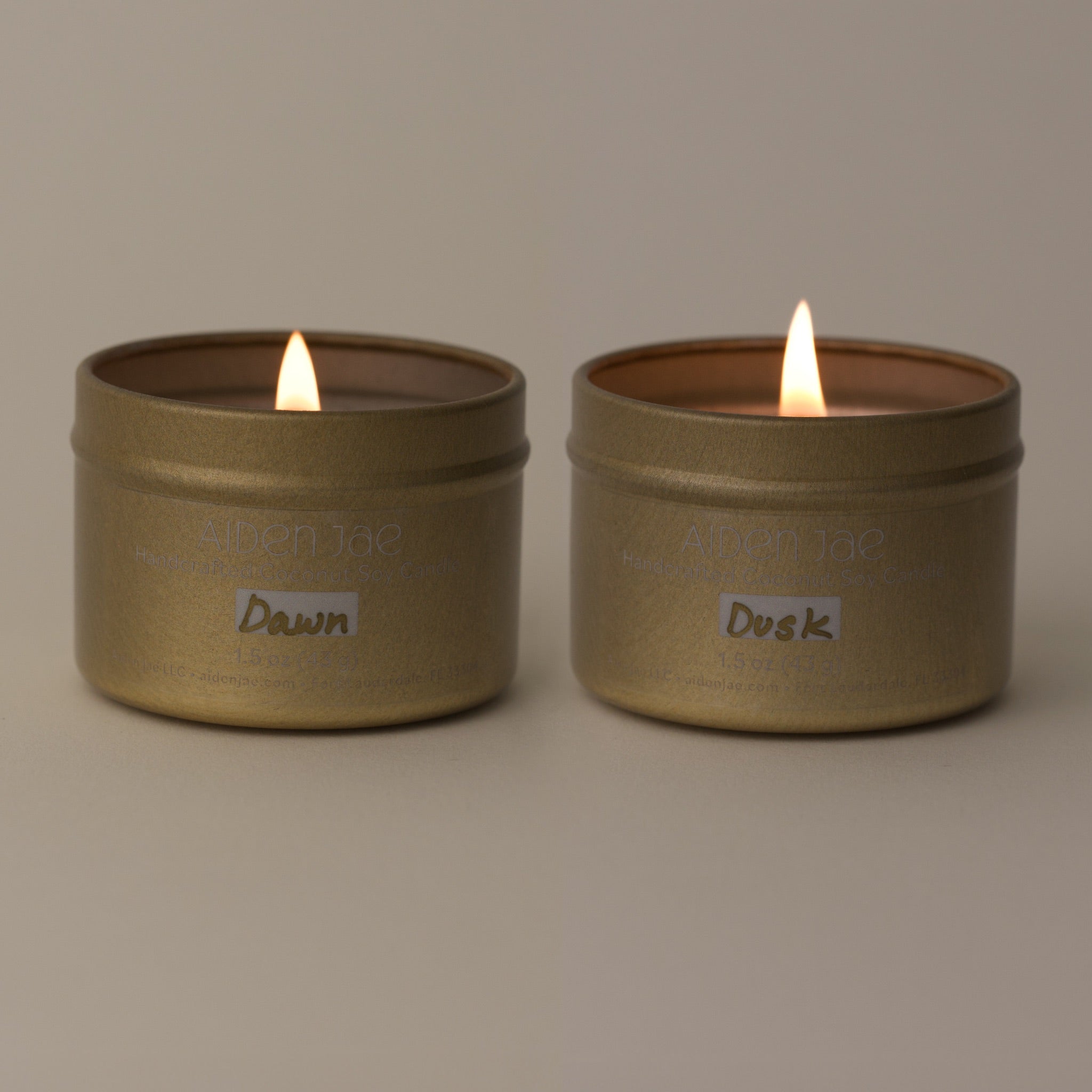 Set of two candles in gold tins, lit with visible flames, in front of a softly dimmed background.