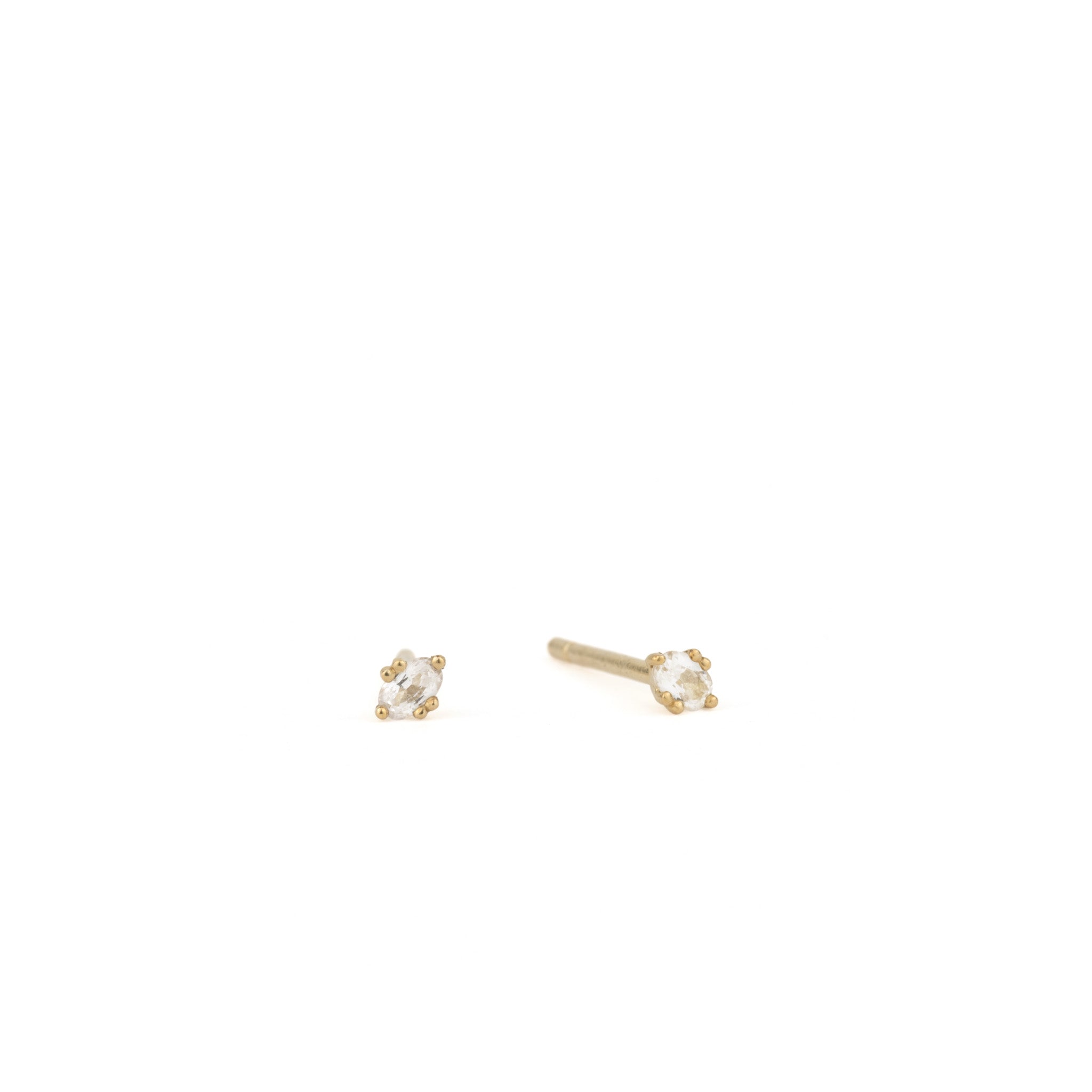 Tiny solid gold oval stud earrings in white sapphire with minimalist design and unique prong setting, on white background.