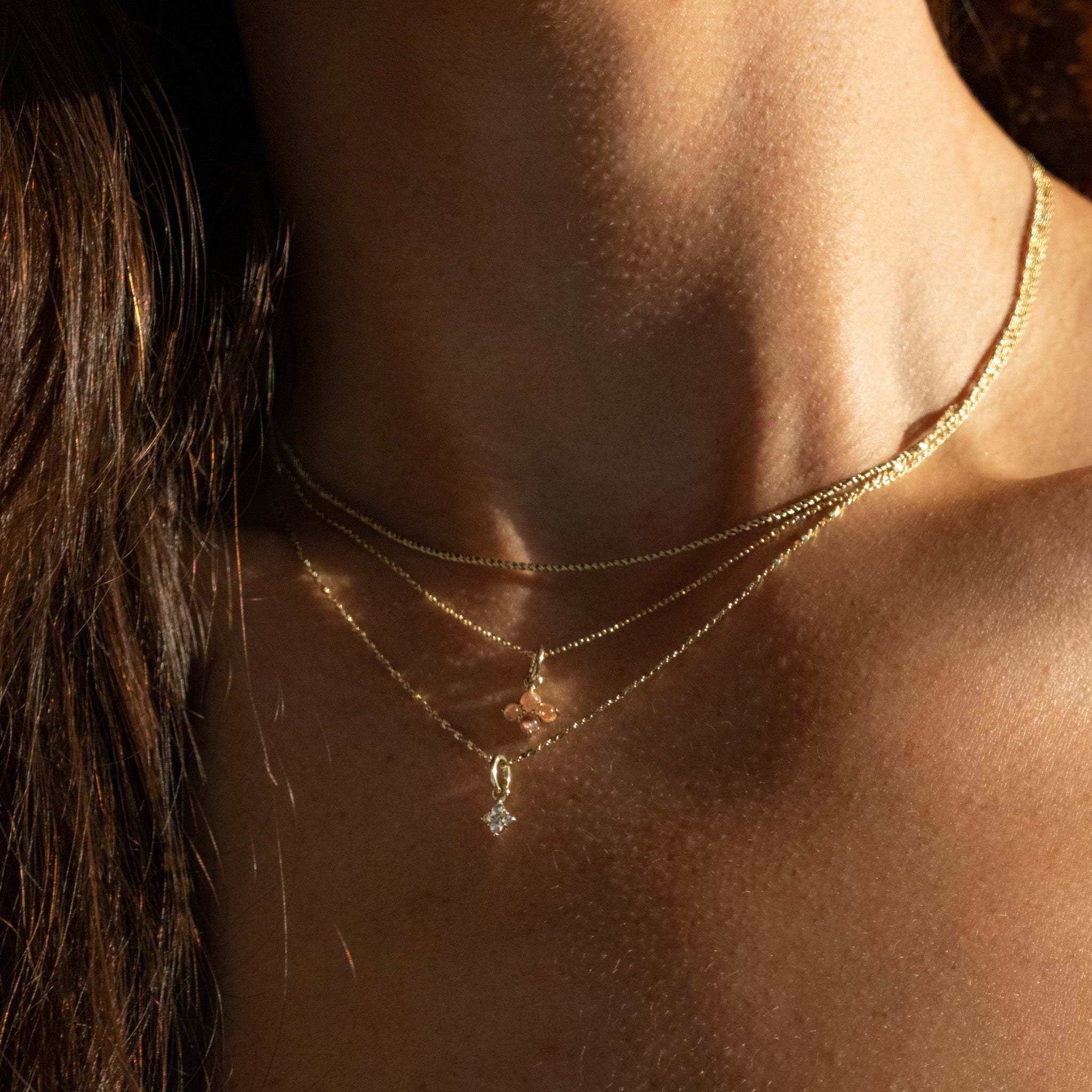 Minimalist solid gold sunstone and white sapphire charm pendants on chain necklaces, layered on neck, glowing in sunlight.