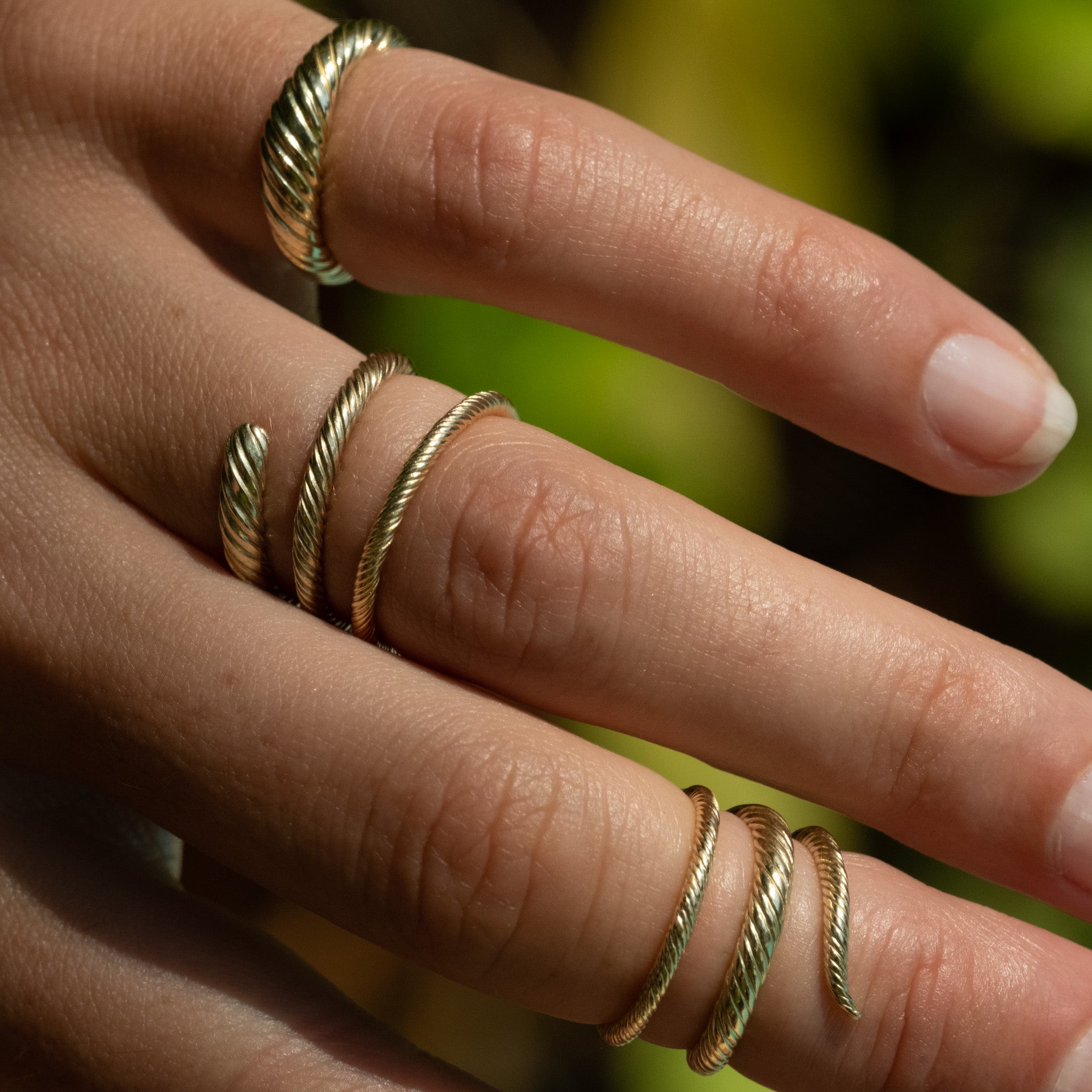 A woman's hand wearing two Aiden Jae Banyan Wrap Rings and an Aiden Jae Dome ring in yellow gold with twisted texture..