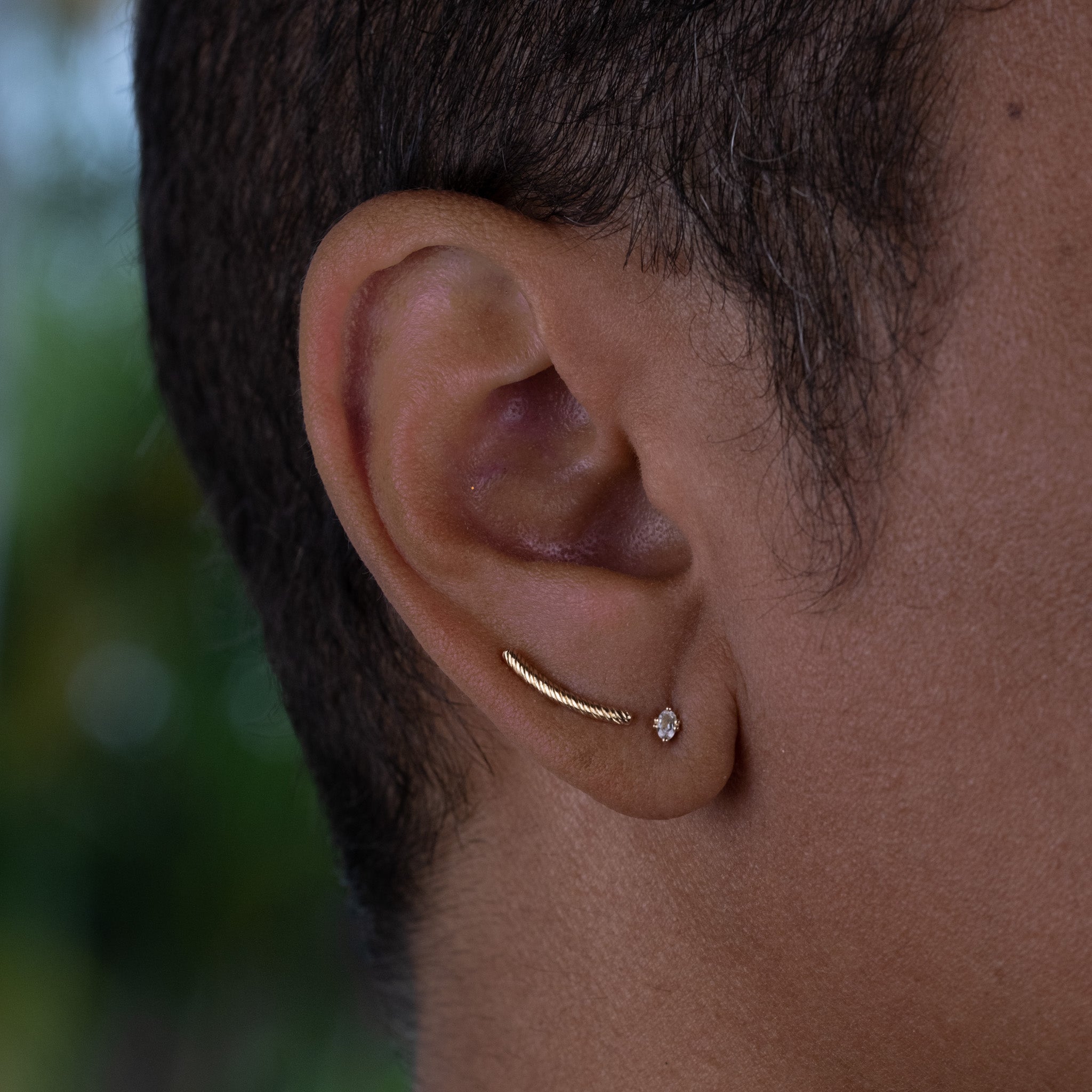 Solid gold bar stud earring with twisted texture and oval white sapphire stud earring worn on ear in a minimalist ear stack.