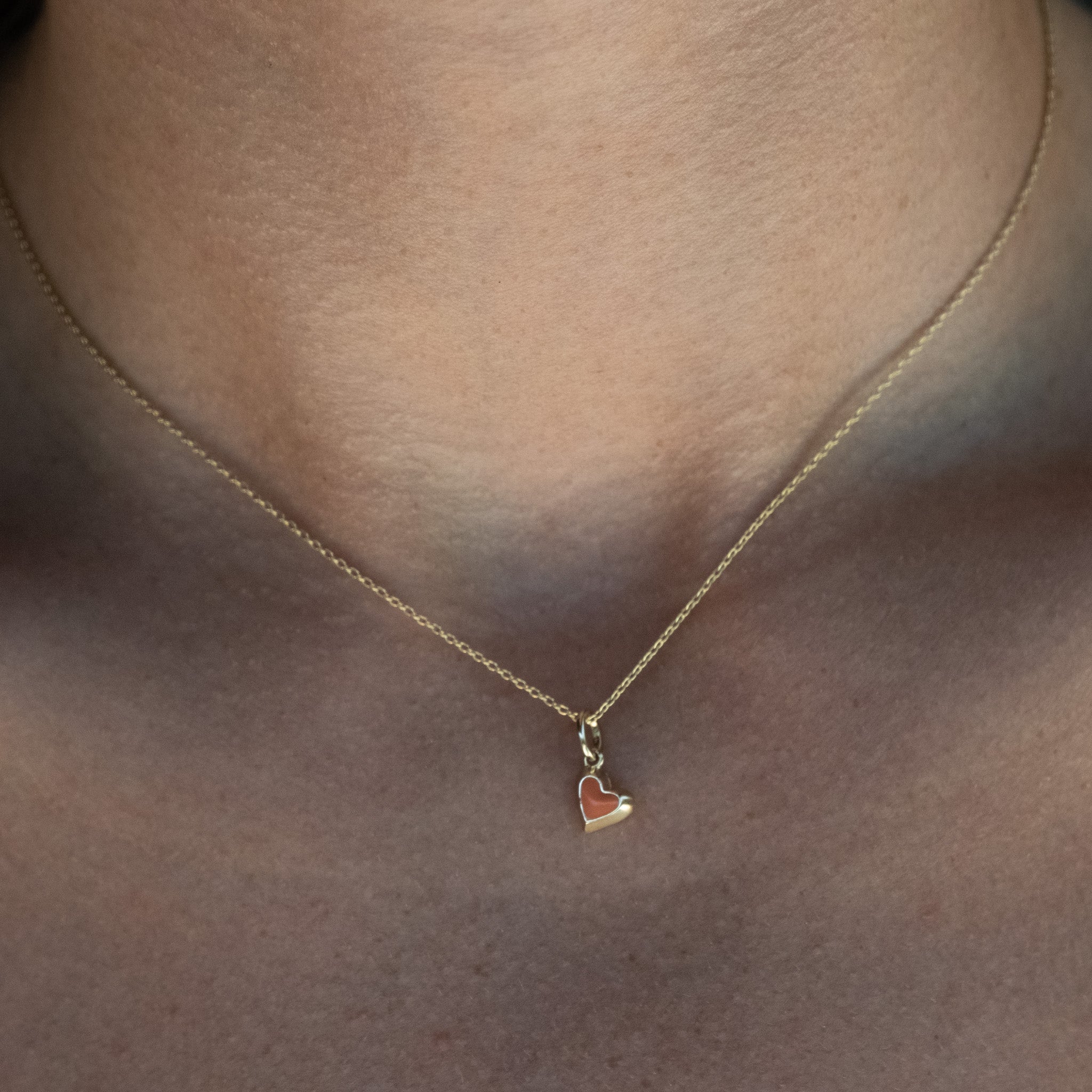 Solid gold tiny heart charm pendant with coral enamel on cable chain, in a minimalist asymmetrical design, worn on neck.