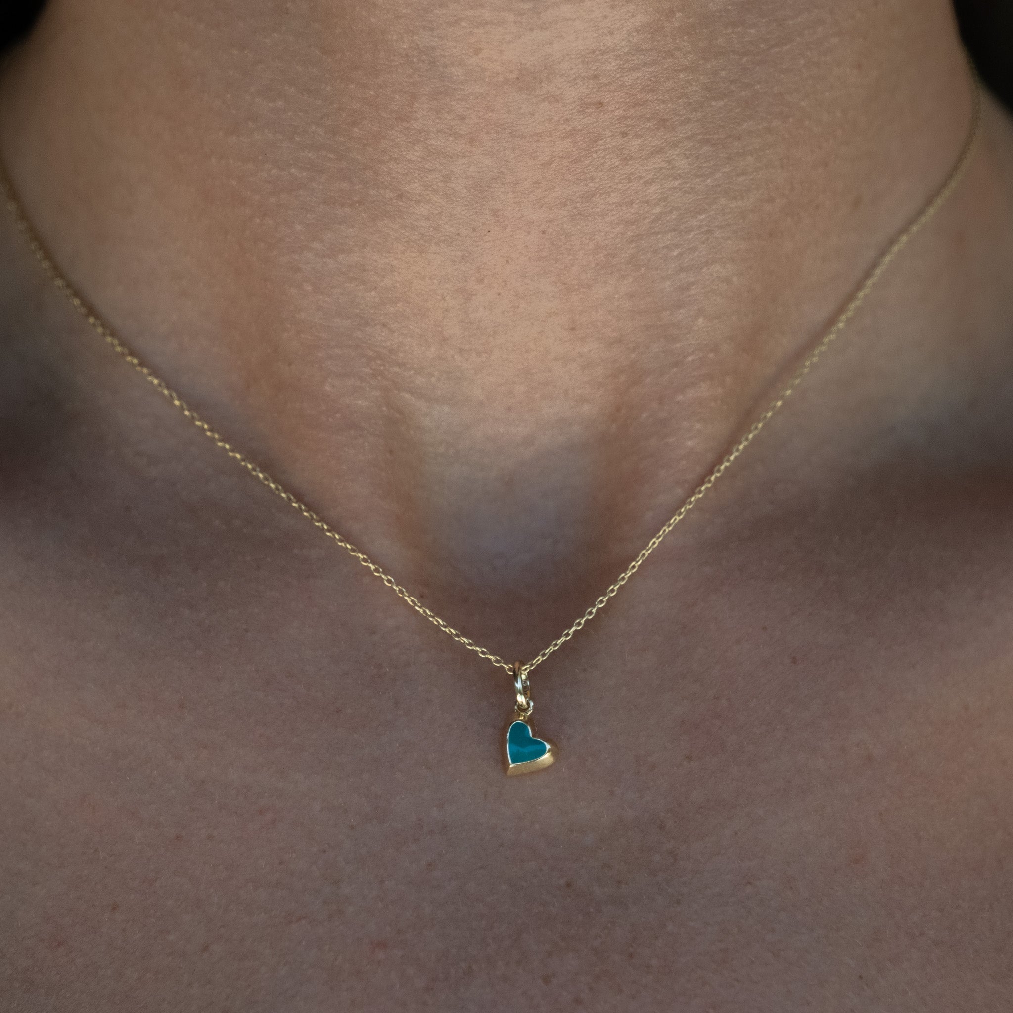 Solid gold tiny heart charm pendant with teal enamel on cable chain, in a minimalist asymmetrical design, worn on neck.