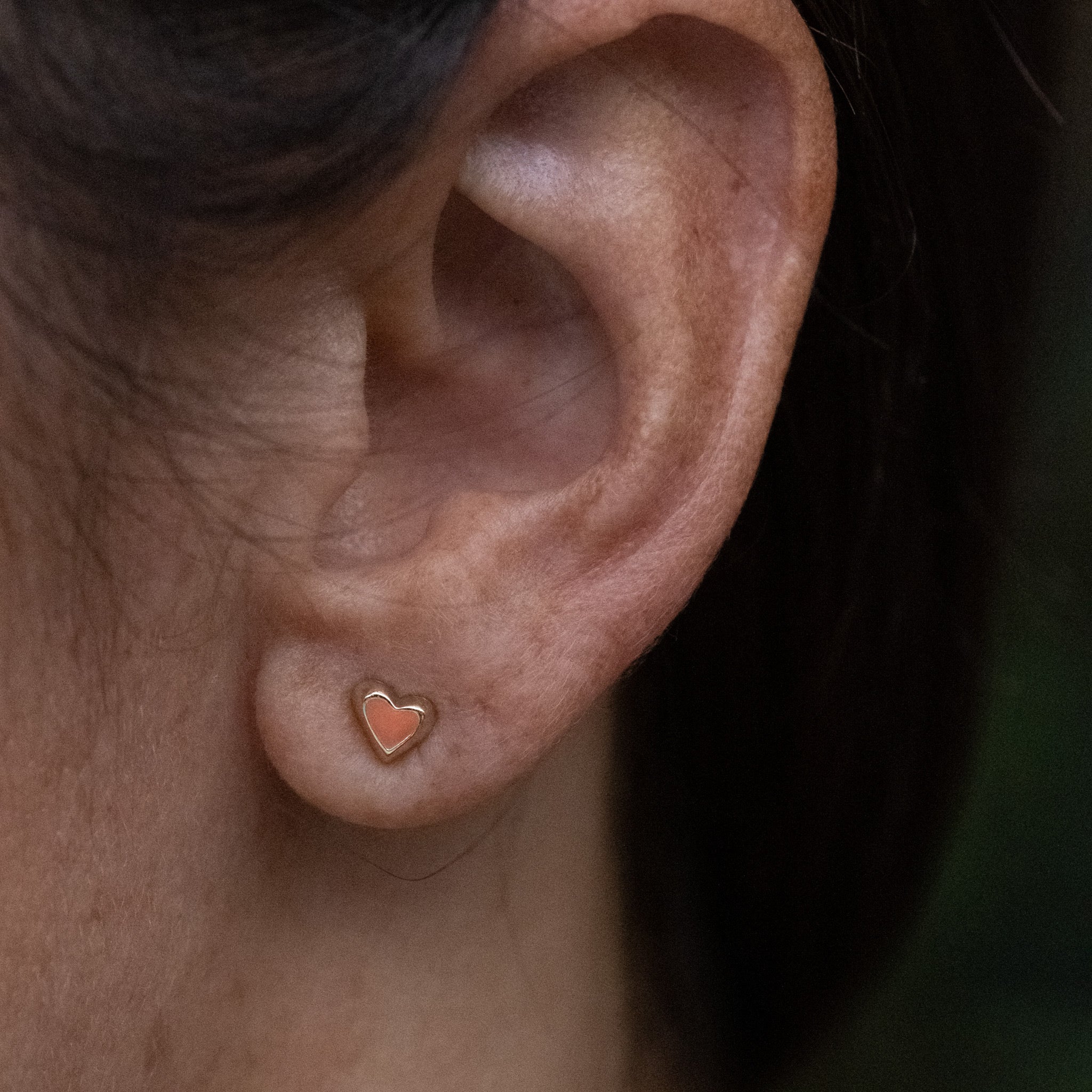 Solid gold tiny heart stud earring with coral enamel, in a minimalist asymmetrical design, worn on ear.