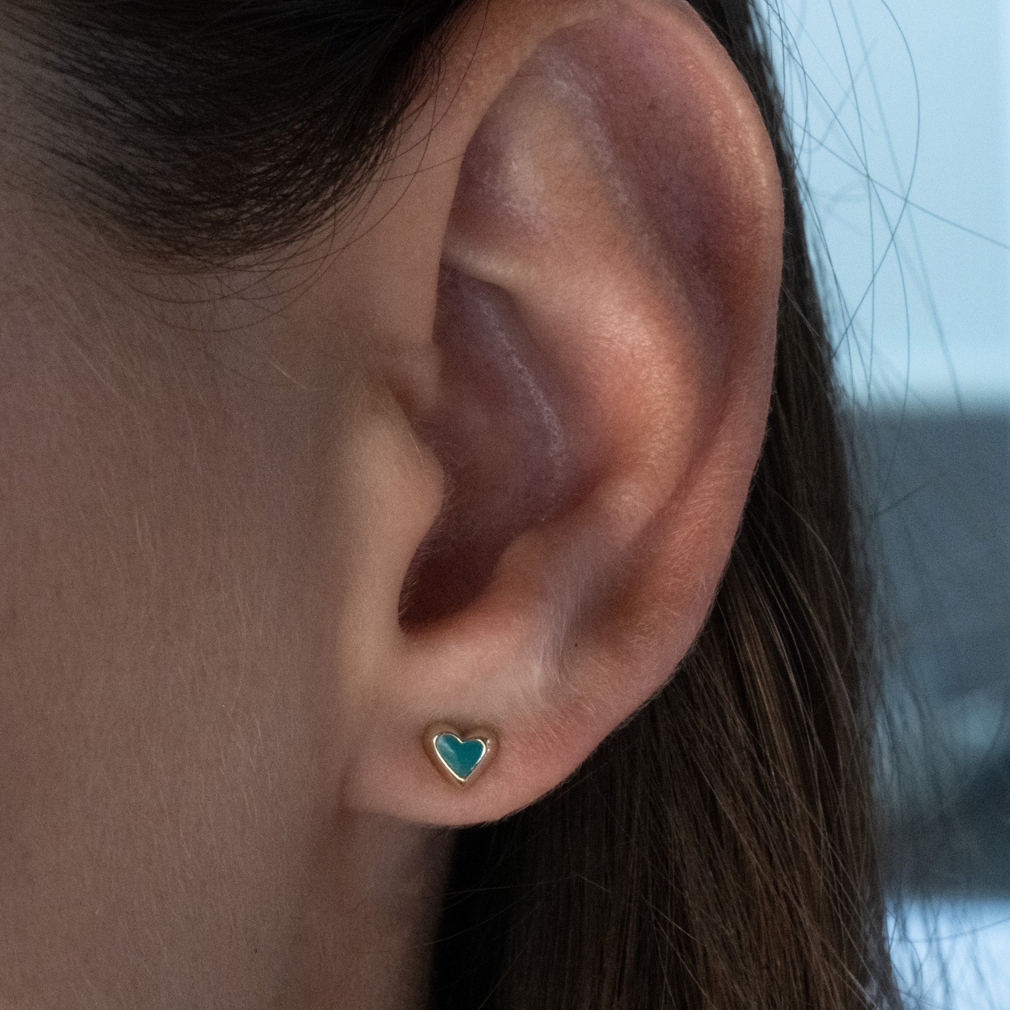Solid gold tiny heart stud earring with teal enamel, worn on ear.
