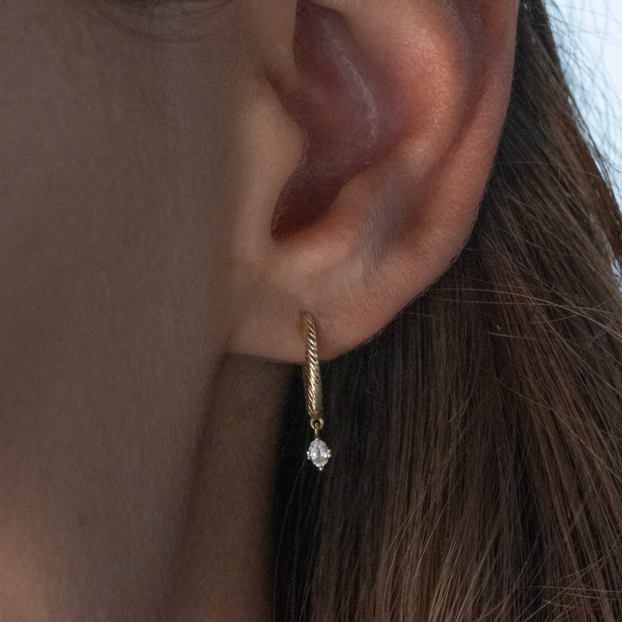 Solid gold huggie hoop earring with twisted texture and dangling oval white sapphire, in minimalist design, worn on ear.