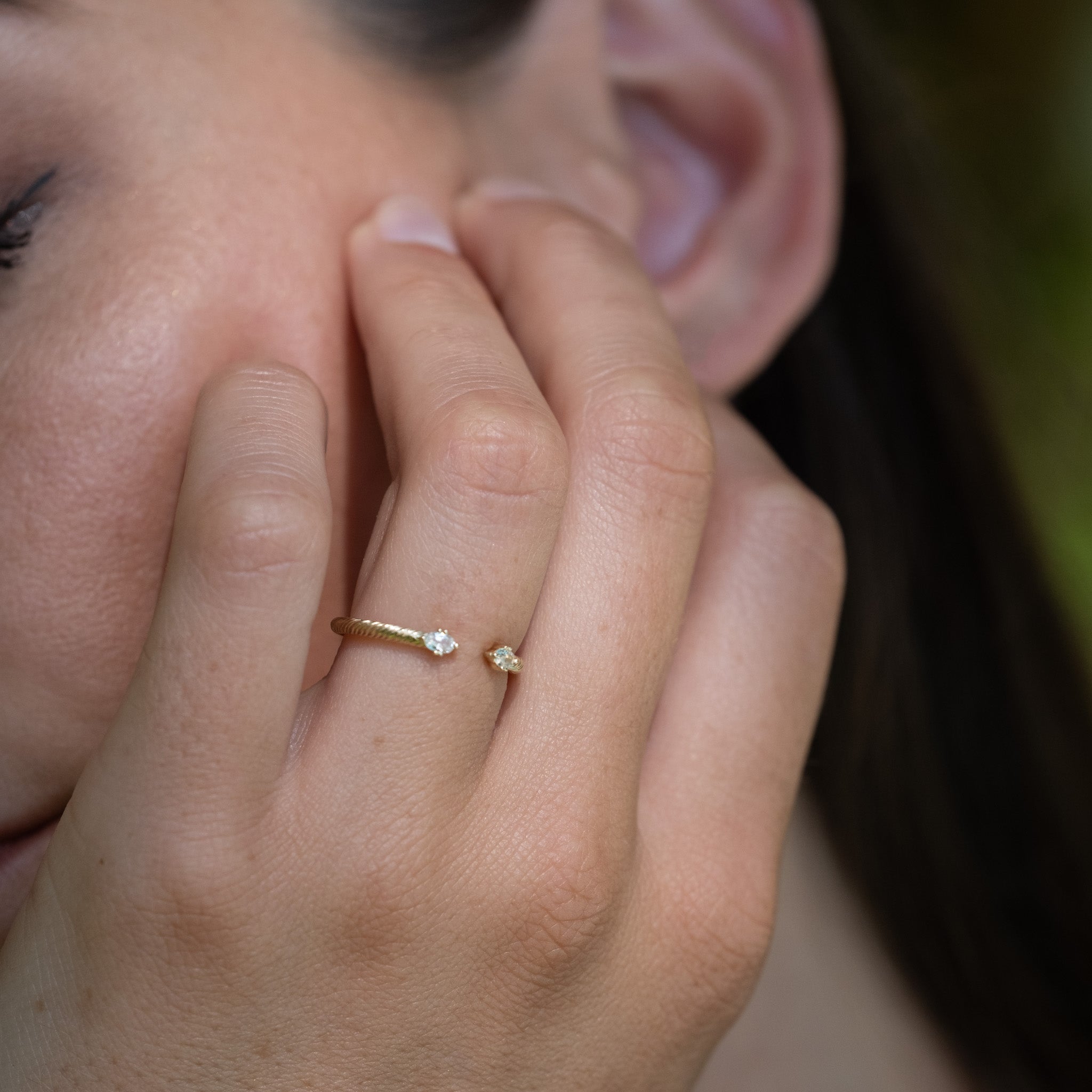 Solid gold stackable open ring with twisted texture and oval white sapphires on ends, shown on hand resting on face.