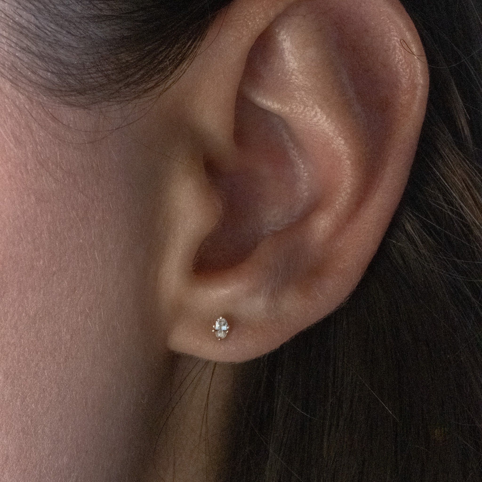 Tiny solid gold oval stud earring in white sapphire with minimalist design and unique prong setting, worn on ear.